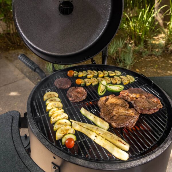 Una parrilla abierta de Pit Boss con carne y vegetales cocinándose, rodeada de un ambiente natural.