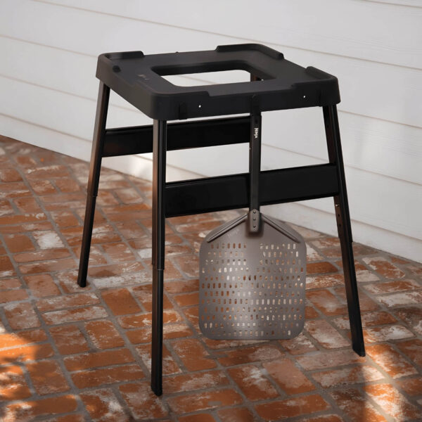 Soporte ajustable negro para hornos exteriores Ninja Woodfire, con diseño robusto, patas reforzadas y espátula metálica colgada en un gancho lateral.