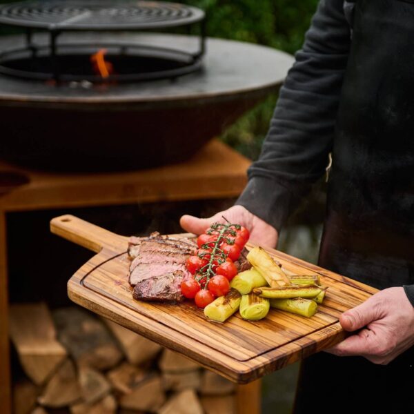 Persona sosteniendo una tabla de madera de teca con carne, tomates cherry y vegetales asados frente a una parrilla