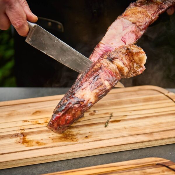 Mano cortando una pieza de carne asada sobre una tabla de madera de teca