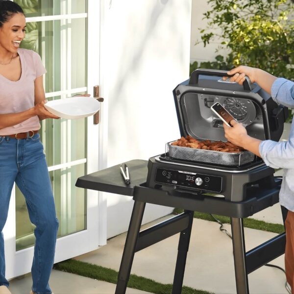 Mujer sosteniendo un plato mientras otra persona prepara alitas de pollo en una barbacoa Ninja Woodfire colocada sobre un soporte ajustable.