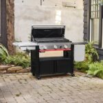 Plancha Weber SLATE GPSB 76 cm Premium con tapa abierta, ubicada en un patio, con diseño moderno, superficie de cocción amplia y almacenamiento inferior.