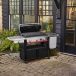 Plancha Weber SLATE GPSB 76 cm Premium con tapa abierta, ubicada en un patio exterior rodeado de plantas, destacando su diseño moderno y funcional.