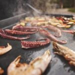 Pulpo, camarones y verduras asándose en la superficie antiadherente de una plancha, con vapor visible durante la cocción.