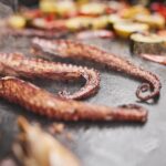 Tentáculos de pulpo dorándose en una plancha junto a verduras frescas, mostrando una textura crujiente y un cocinado uniforme.