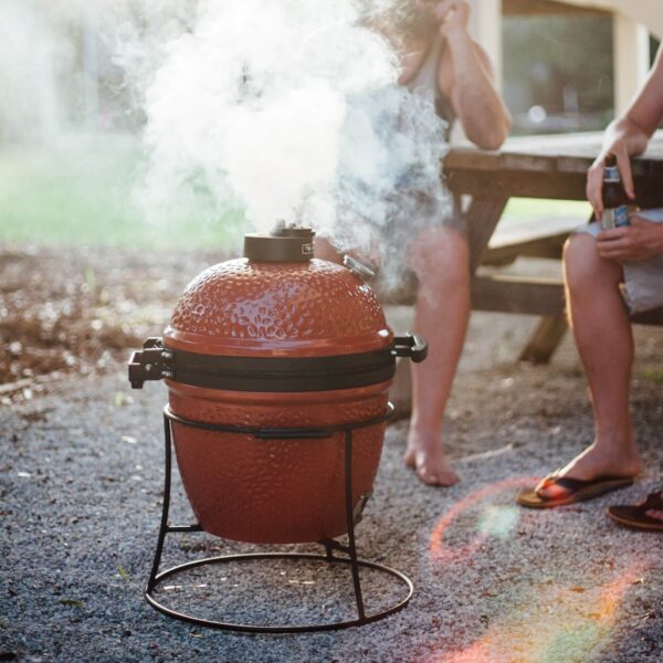 Kamado Joe Jr. emitiendo humo mientras se cocina al aire libre, con personas disfrutando alrededor.