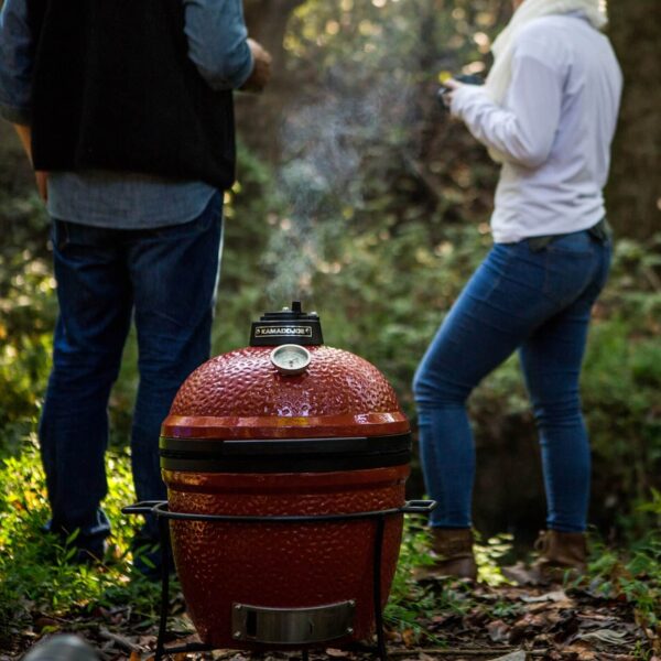 Kamado Joe Jr. emitiendo humo con dos personas en el fondo disfrutando de la naturaleza.