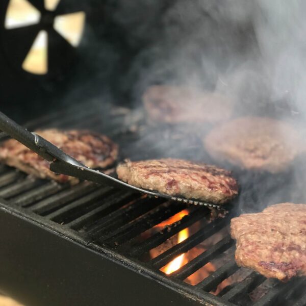 Hamburguesas cocinándose en las rejillas de la Caja Portátil de Cocción Lateral CharGriller, con humo y fuego visible.