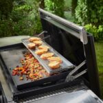 Soporte calentador de dos niveles en una plancha Weber de 76 cm, utilizado para mantener pan tostado caliente mientras se cocinan vegetales en la superficie principal.