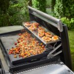 Soporte calentador de dos niveles en uso sobre una plancha Weber de 76 cm, con alimentos como panes y vegetales organizados y cocinados al aire libre.