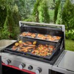 Plancha Weber de 76 cm en uso al aire libre, con un soporte calentador de dos niveles que mantiene panes y guarniciones calientes mientras se cocinan camarones, vegetales y limones en la superficie principal.
