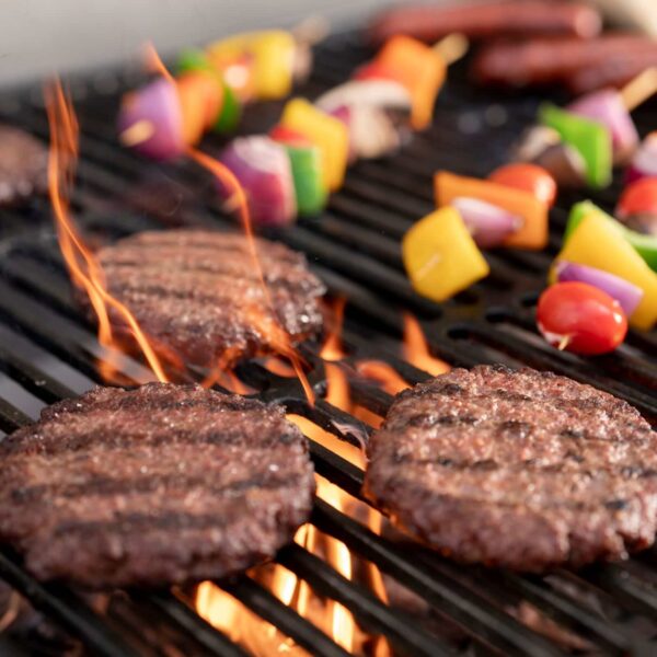 Hamburguesas y brochetas de vegetales asándose en la parrilla CharGriller, con llamas visibles para un sellado perfecto.