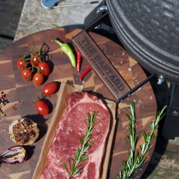 Tabla de cortar The Bastard de madera de acacia con tomates, pimientos, ajo asado y un corte de carne con romero, junto a una parrilla de exterior