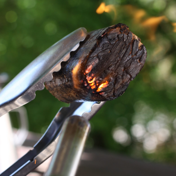 Bloque de madera encendido sujetado con pinzas metálicas, listo para ahumar en BBQ.