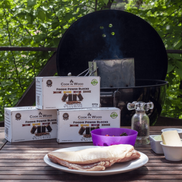 Bloques de madera Cook in Wood junto a ingredientes para BBQ, con parrilla y accesorios listos para ahumar.