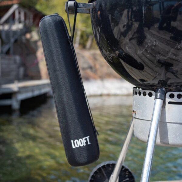 Funda negra de la marca Looft colgada de una barbacoa al aire libre, junto a un paisaje con agua y vegetación.