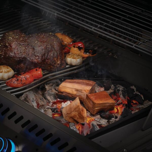 Trozos de madera de manzano en las brasas de una parrilla de gas, ahumando un asado grande junto con verduras.