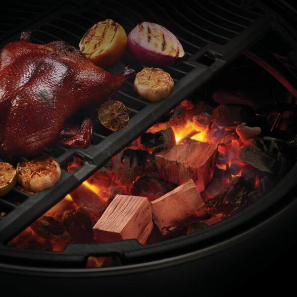 Trozos de madera de cerezo incandescentes bajo un pato asado en una parrilla circular, acompañados de cebollas y limones caramelizados.