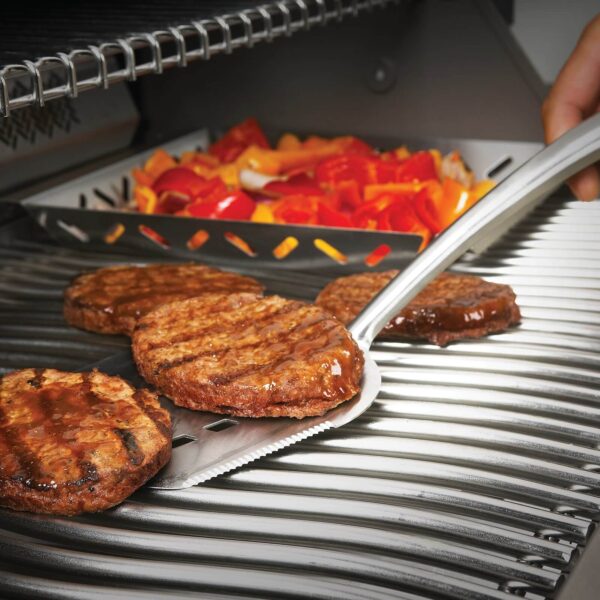 Espátula de acero inoxidable manejando una hamburguesa en una parrilla, con vegetales asados al fondo.