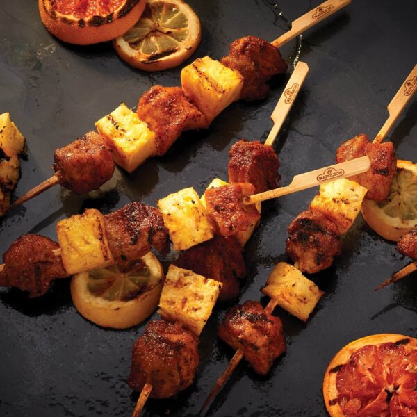 Brochetas de carne y piña ensartadas en pinchos de bambú de 30,5 cm sobre una superficie oscura, acompañadas de rodajas de limón asado.