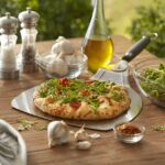 Pala para pizza de acero inoxidable Weber con una pizza recién hecha sobre una mesa, rodeada de ingredientes frescos como ajo, rúcula, aceite de oliva, sal y pimienta