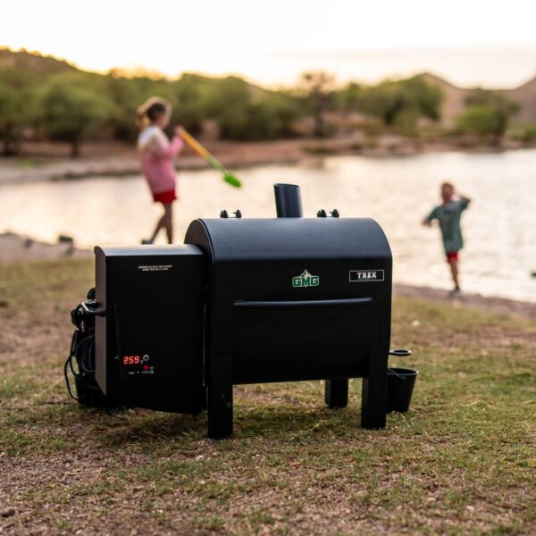 Barbacoa de pellets Trek Prime 2.0 de Green Mountain Grills en un entorno natural junto a un lago, con niños jugando en el fondo y la pantalla digital mostrando 259°F.