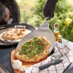 Pala para pizza de acero inoxidable Weber levantando una pizza recién hecha en una barbacoa, con una pizza adicional al fondo y utensilios de cocina alrededor