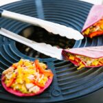 Pinzas de acero inoxidable manipulando tacos en una parrilla con relleno de vegetales y pollo.