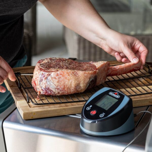 Persona sazonando un corte grueso de carne cerca de la Sonda Digital Accu-Probe Bluetooth sobre una superficie de cocina.