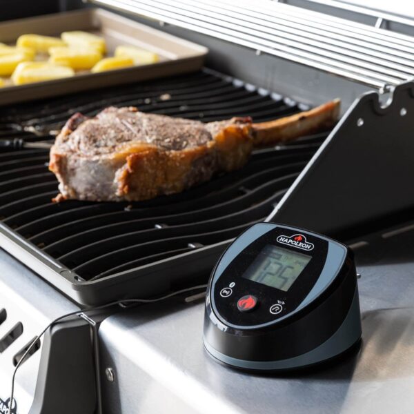 Sonda Digital Accu-Probe Bluetooth mostrando la temperatura mientras se asa un corte de carne con hueso en una parrilla, junto a unas rodajas de patata.