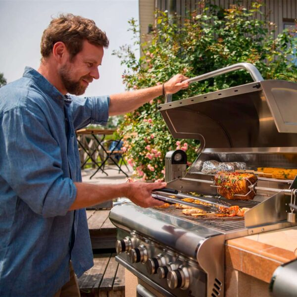 Hombre cocinando en la barbacoa de gas Ardor PTS 5.0 con varios alimentos, incluyendo maíz y carne asada, en un jardín.