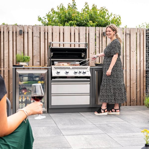 Mujer cocinando en la barbacoa de gas Napoleon Prestige Pro 500 RB en una cocina al aire libre, con otra persona disfrutando de una copa de vino.