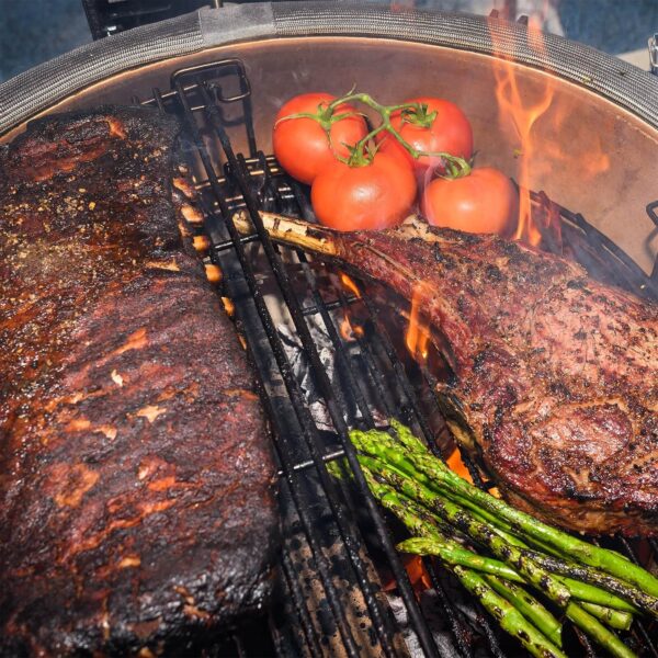 Parrilla del Kamado Big Joe II con diversos alimentos cocinándose, incluyendo carne, tomates y espárragos.