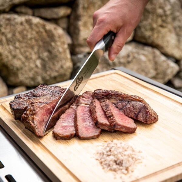 Mano cortando un trozo de carne asada con el Cuchillo Chef Napoleon en una tabla de cortar de madera.