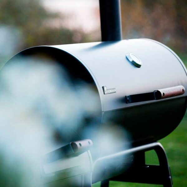 Imagen atmosférica de la barbacoa Black Taurus 660+ en uso, con humo aromático ascendiendo en un entorno al aire libre.