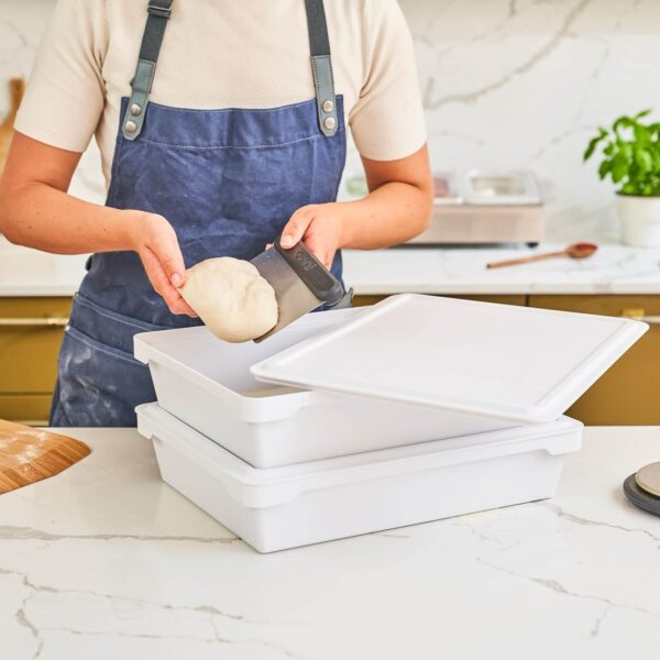 Una persona en delantal azul preparando masa de pizza y utilizando un raspador de masa sobre unas cajas de almacenamiento de plástico blanco en una cocina clara.