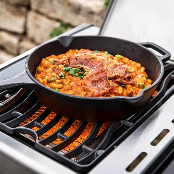 Guiso casero de frijoles con trozos de carne en una sartén de hierro fundido, cocinándose en una parrilla al aire libre.