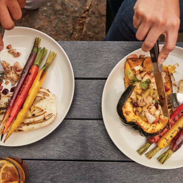 Manos de una persona cortando vegetales asados coloridos servidos en platos sobre una mesa de madera, resultado de cocinar en la barbacoa Kettle Ø 57cm 2024.