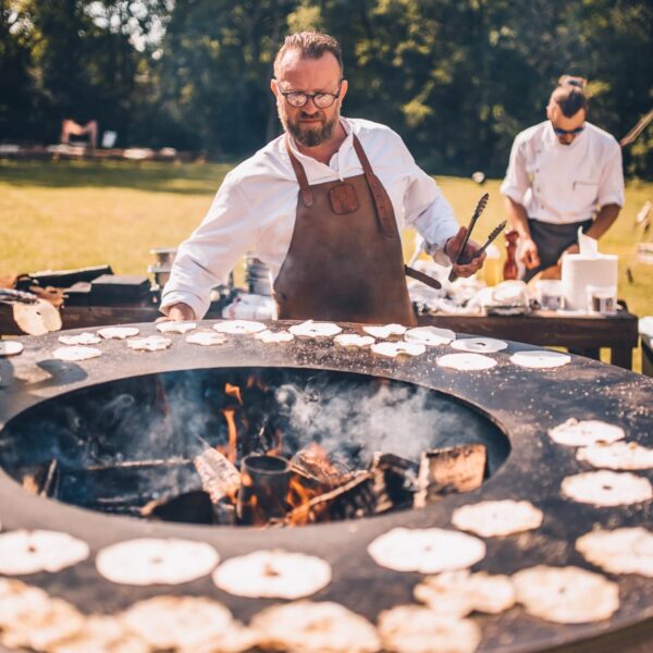 Chef cocinando en una barbacoa de leña circular durante un evento al aire libre, con tortillas cocinándose en la superficie de la parrilla.