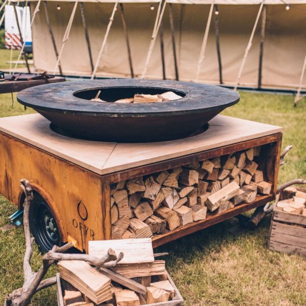 Barbacoa de leña circular sobre un remolque con almacenamiento integrado de madera en un evento al aire libre.