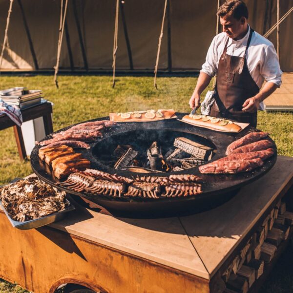 Chef asando una variedad de carnes y filetes de salmón en una barbacoa de leña circular durante un evento al aire libre.