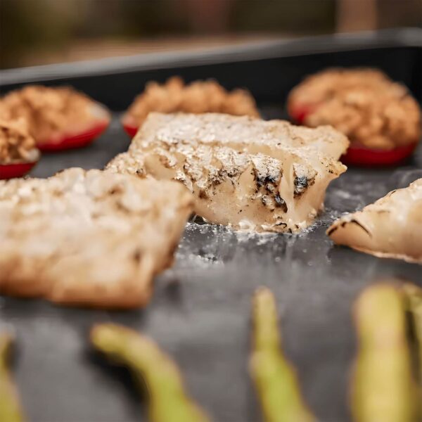 Primer plano de filetes de pescado cocinándose en la plancha Weber SLATE GP 56 cm Premium, con pimientos rellenos y espárragos en el fondo.