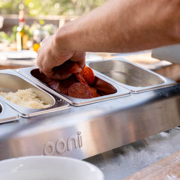 Mano de chef tomando pepperoni de un envase de acero inoxidable Ooni, destacando ingredientes frescos y la calidad de la herramienta de cocina.