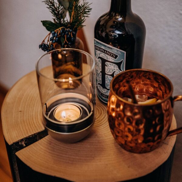 Vela Gravity de Höfats encendida sobre una mesa de madera junto a una botella de licor y una taza de cobre