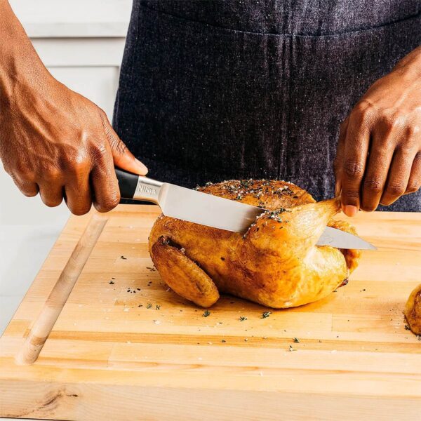 Persona cortando un pollo asado con un cuchillo de cocina Ninja sobre una tabla de cortar de madera. El cuchillo se usa para separar la pierna del resto del pollo, mostrando su precisión y filo.