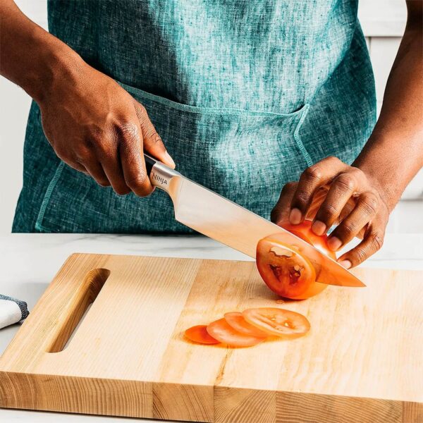 Persona cortando un tomate con un cuchillo de cocina sobre una tabla de madera. El cuchillo tiene la marca "Ninja" y el mango ergonómico. Se ve un delantal azul y una toalla de cocina a un lado.