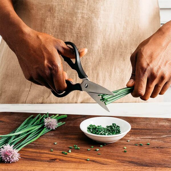 Persona usando tijeras de cocina para cortar cebollino sobre una tabla de madera. El cebollino cortado se coloca en un pequeño plato blanco.