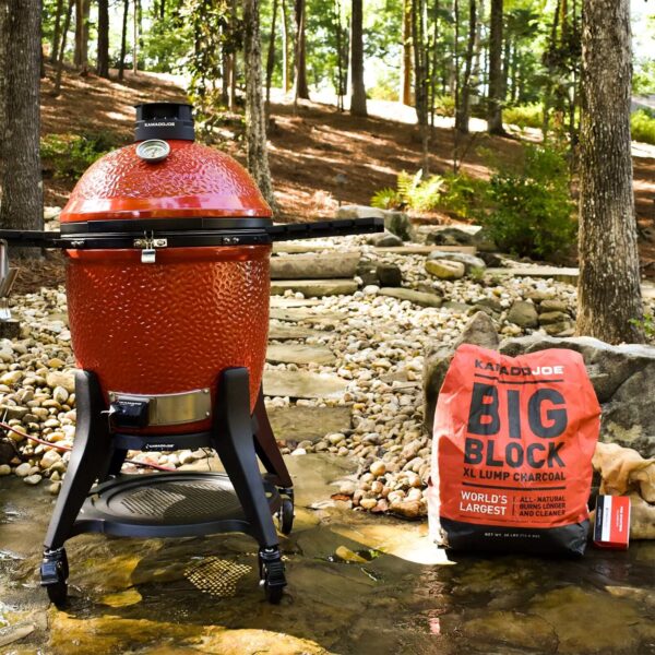 Parrilla Kamado Joe junto a saco de carbón Big Block en un entorno natural al aire libre.