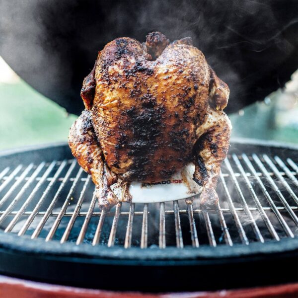 Pollo asado sobre el soporte Kamado Joe en una parrilla, con humo alrededor, destacando la cocción uniforme y la piel crujiente.