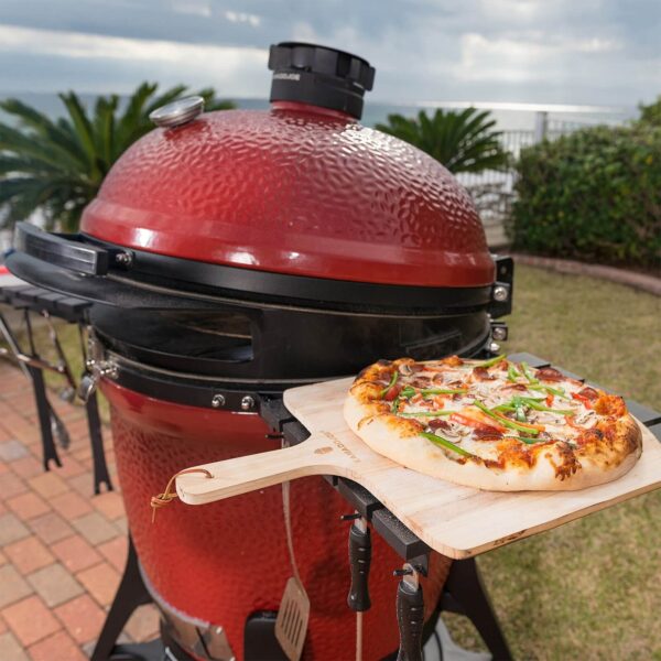 Pala de pizza Kamado Joe con una pizza recién horneada frente a una parrilla Kamado Joe roja.